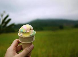 feche uma mão segurando sorvete de cor de arco-íris em cone de waffle, fundo desfocado de campo de arroz tropical verde e montanha foto