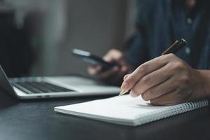 mão de homem está escrevendo em um bloco de notas com uma caneta e um telefone inteligente na mesa. foto
