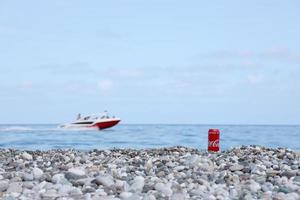 antalya, turquia - 18 de maio de 2021 lata vermelha original da coca cola encontra-se em pequenas pedras redondas perto da costa do mar. lata de coca-cola e lancha na praia foto