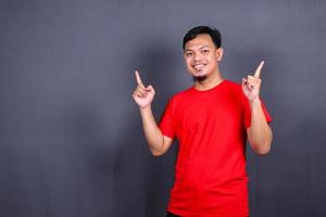 jovem asiático em camiseta vermelha apontando com o dedo isolado no fundo cinza foto