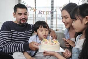 feliz família tailandesa asiática, filho jovem é surpreendido com bolo de aniversário, sopra a vela e celebra a festa da alegria com os pais e a irmã na sala de estar juntos, evento especial doméstico de bem-estar. foto