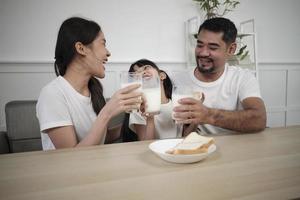 uma família tailandesa asiática saudável, uma filha pequena e pais jovens bebem leite branco fresco em vidro e pão alegria juntos em uma mesa de jantar de manhã, bem-estar nutrição casa café da manhã refeição estilo de vida. foto