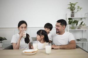 uma família tailandesa asiática saudável, crianças pequenas e pais jovens bebem leite branco fresco em vidro e pão alegria juntos em uma mesa de jantar de manhã, bem-estar nutrição em casa café da manhã refeição estilo de vida. foto