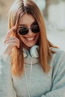 uma garota moderna com óculos de sol andando na rua ouve música em fones de ouvido. foto
