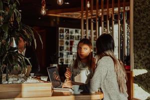 duas mulheres de negócios jovens sentados à mesa no café. garota mostra informações do colega na tela do laptop. garota usando smartphone, blogando. trabalho em equipe, reunião de negócios.. foto