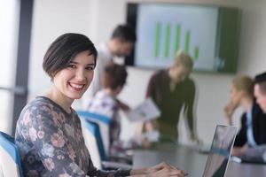 jovem mulher de negócios no escritório trabalhando no laptop com equipe na reunião em segundo plano foto