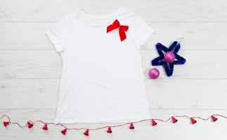 feche a camiseta branca modelo em branco com espaço de cópia e conceito de férias de natal. t-shirt de maquete de vista superior e laço vermelho sobre fundo branco de madeira. feliz ano novo acessórios de decorações. roupa de natal foto