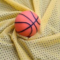 pequena bola de basquete de borracha laranja encontra-se em uma textura de tecido de roupa de camisa esportiva amarela e fundo com muitas dobras foto