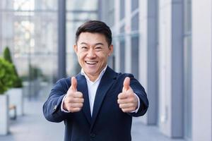 corretor empresário asiático em terno de negócios feliz olhando para a câmera e mostrando os polegares foto