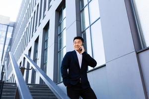 empresário asiático de sucesso falando ao telefone perto do centro de escritórios sorrindo foto