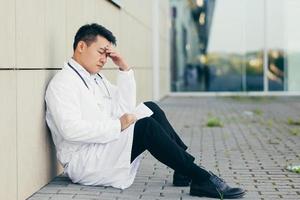 retrato médico homem asiático cansado depois do trabalho sentado no chão perto da clínica desapontado foto