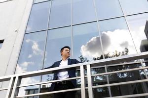 chefe de empresário asiático fica perto de seu centro de escritório na varanda em um terno de cor escura foto