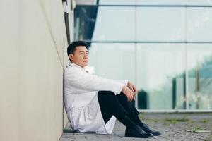 retrato médico homem asiático cansado depois do trabalho sentado no chão perto da clínica desapontado foto