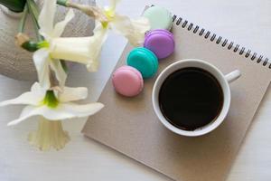 bloco de notas, macarons, xícara de café e narcisos em um vaso em uma mesa branca. local de trabalho inspirador. foto