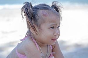 adorável menina criança feliz brincando com areia na praia do oceano durante as férias tropicais de verão. cara engraçada criança olhando mãe com grande marca de nascença vermelha de hemangiomas capilares de morango na cabeça foto