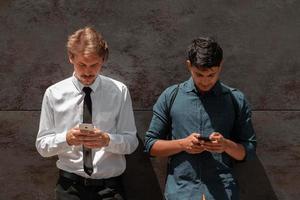 homens de negócios de inicialização multiétnica casual, um deles é indiano usando smartphone durante o intervalo do trabalho em frente à parede cinza do lado de fora foto