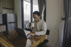 um jovem asiático passa seu tempo em casa sentado na sala de jantar chocado enquanto trabalha em seu laptop enquanto segura uma caneca foto