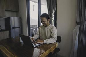 um jovem asiático passa seu tempo em casa sentado na sala de jantar trabalhando alegremente em seu laptop foto