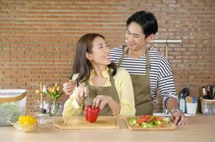 jovem casal asiático sorridente vestindo um avental na sala da cozinha, cozinhando o conceito foto
