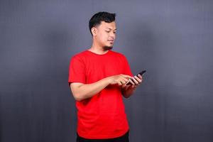retrato de um jovem asiático alegre vestindo camiseta vermelha em pé isolado sobre fundo cinza, usando telefone celular foto