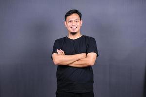 sorrindo bonito homem asiático em pé de t-shirt preta com os braços cruzados isolados no fundo cinza foto