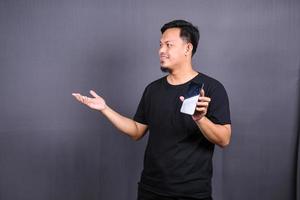 foto de cara asiático atraente segurando o telefone celular vestindo camiseta preta isolada fundo de cor cinza