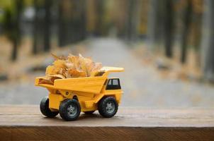 um pequeno caminhão amarelo de brinquedo é carregado com folhas amarelas caídas. o carro fica em uma superfície de madeira contra um fundo de um parque de outono embaçado. limpeza e remoção de folhas caídas. trabalhos sazonais foto