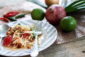 salada de mamão tailandês em um prato, salada de mamão com cocô, limão, pimenta, tomate vermelho em tigela branca na mesa de madeira foto