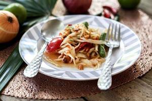 salada de mamão tailandês em um prato, salada de mamão com cocô, limão, pimenta, tomate vermelho em tigela branca na mesa de madeira foto