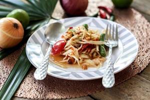 salada de mamão tailandês em um prato, salada de mamão com cocô, limão, pimenta, tomate vermelho em tigela branca na mesa de madeira foto