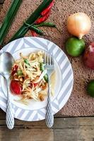 salada de mamão tailandês em um prato, salada de mamão com cocô, limão, pimenta, tomate vermelho em tigela branca na mesa de madeira foto