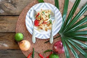 salada de mamão tailandês em um prato, salada de mamão com cocô, limão, pimenta, tomate vermelho em tigela branca na mesa de madeira foto
