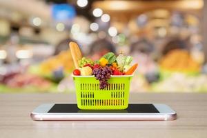 alimentos frescos e legumes na cesta de compras no smartphone móvel na mesa de madeira com o conceito on-line de mercearia de fundo desfocado do corredor do supermercado foto