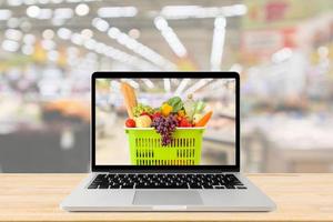 corredor de supermercado fundo desfocado com computador portátil e cesta de compras no conceito on-line de mercearia de mesa de madeira foto