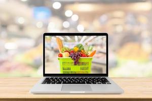 corredor de supermercado fundo desfocado com computador portátil e cesta de compras no conceito on-line de mercearia de mesa de madeira foto