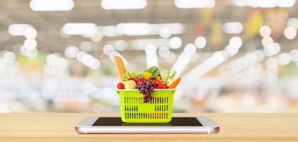 alimentos frescos e legumes na cesta de compras no smartphone móvel na mesa de madeira com o conceito on-line de mercearia de fundo desfocado do corredor do supermercado foto