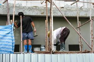 equipe de dois trabalhadores fica em andaimes de madeira instalam uso para alto nível. cimento de gesso de parede de concreto na parede para construir uma nova casa. foto