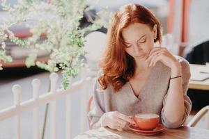 mulher europeia ruiva encantadora bebe cappuccino ou café no café do terraço, tem expressão facial calma, usa suéter marrom, tem maquiagem, gosta de uma boa bebida. conceito de pessoas e estilo de vida. foto