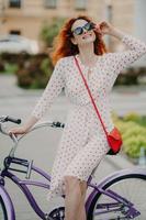 mulher alegre ativa anda de bicicleta na cidade, usa óculos de sol elegantes, tem um sorriso encantador, vestido de verão, aproveita o dia ensolarado, passa o tempo livre ativamente. pessoas, transporte e estilo de vida foto