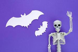 esqueleto com dois morcegos voadores cortados em papel branco como bolhas de fala em branco sobre fundo violeta de halloween. modelo de plástico anatômico esqueleto humano com uma mão levantada afasta pensamentos obsessivos. foto