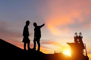 silhueta de engenheiro e trabalhador verificando projeto no fundo do canteiro de obras, canteiro de obras de infraestrutura ao pôr do sol na noite foto