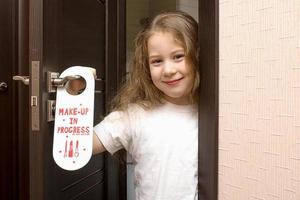 uma garotinha com lábios pintados espreita por trás de uma porta que diz não perturbe a maquiagem em andamento foto