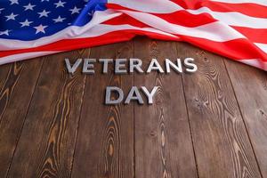 o dia dos veteranos de palavras colocado com letras de metal prateado na superfície da placa de madeira com bandeira dos eua amassada foto