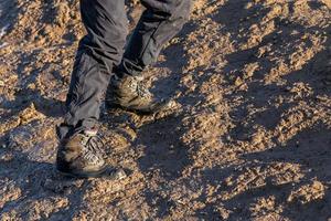 pernas em calças cinzentas e botas de trekking subindo na colina lamacenta à luz do sol da noite foto