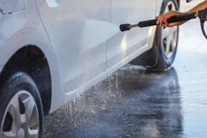 mão com lavadora de alta pressão lavando carro branco na estação de lavagem de carros de autoatendimento público foto