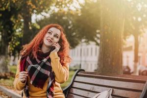 jovem ruiva de moletom amarelo, falando no telefone. senhora senta-se no banco do parque no dia do outono. estilo de vida. foto