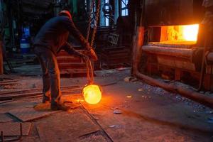 operário de forja movendo a peça de trabalho de metal quente brilhante com focreps de guindaste de corrente foto