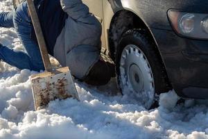 homem trabalhando no carro preso na neve no joelho com pá à luz do dia offroad foto