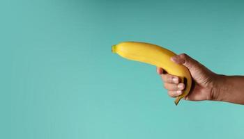 imagem do conceito de banana. mão segurando uma banana contra o fundo azul, parece uma arma. foto metáfora. estilo limpo e minimalista