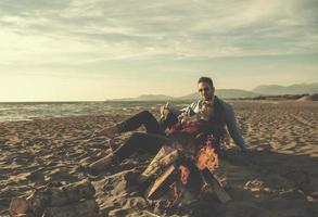 jovem casal sentado na praia ao lado da fogueira bebendo cerveja foto